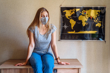 Beautiful girl stays at home because of coronovirus, sitting near the world map and dreaming of travels after the epidemic is over and all the sick will recover. Isolated at home in a protective mask.の写真素材