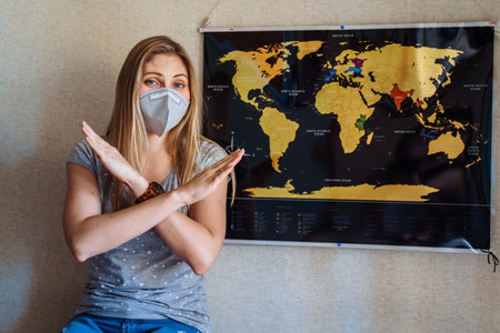 Blond girl in a protective mask staying at home because of coronovirus, sitting near the world map and showing stop sign about journeys and traveling during epidemyの写真素材