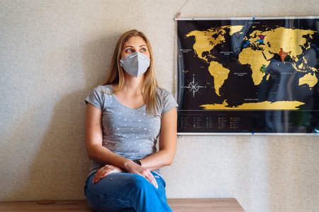 Beautiful girl stays at home because of coronovirus, sitting near the world map and dreaming of travels after the epidemic is over and all the sick will recover. Isolated at home in a protective mask.の写真素材