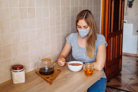 Attractive blonde is sitting in the kitchen near the table with food. She is isolated at home during quarantine due to the  epidemic, wearing a protective mask.Looking to plate with soup.の写真素材
