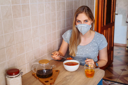 Economic blonde woman cooked red soup and eats it while staying isolated at home on quarantine. She wears a protective face mask to reduce the chance of getting sick during a  epidemic.の写真素材