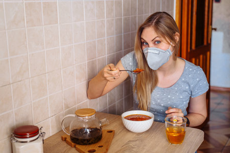 Economic blonde woman cooked red soup and eats it while staying isolated at home on quarantine. She wears a protective face mask to reduce the chance of getting sick during a  epidemic.の写真素材