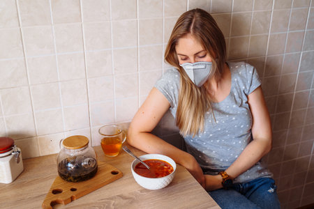Attractive blonde is sitting in the kitchen near the table with food. She is isolated at home during quarantine due to the  epidemic, wearing a protective mask.Looking to plate with soup.の写真素材