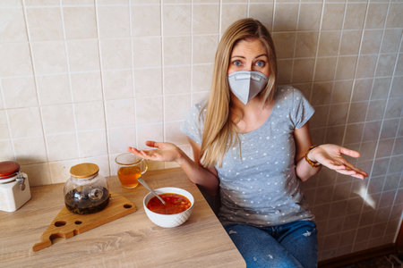 Attractive blonde is sitting in the kitchen near the table with food. She is isolated at home during quarantine due to the  epidemic, wearing a protective mask.Looking to plate with soup.の写真素材