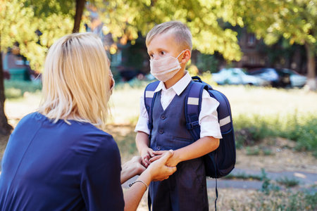 Mom is ready to send her little son to first grade. Gently holds the boy's hands. They are wearing protective medical masks. New requirements for training during the quarantine periodの写真素材