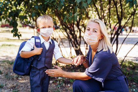 Mom is ready to send her little son to first grade. Gently holds the boy's hands. They are wearing protective medical masks. New requirements for training during the quarantine periodの写真素材