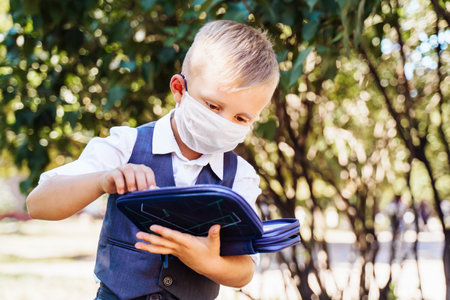 Young student of first grade stands in the schoolyard. Took out pencil from the pencil case. Wearing protective medical mask due to new safety requirements duringの写真素材