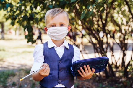Young student of first grade stands in the schoolyard. Took out pencil from the pencil case. Wearing protective medical mask due to new safety requirementsの写真素材
