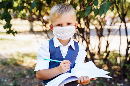 Young student of first grade stands in the schoolyard. Took out pencil from the pencil case. Wearing protective medical mask due to new safety requirementsの写真素材
