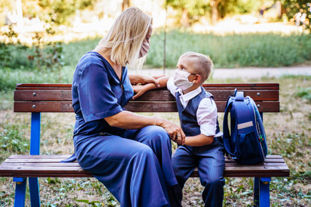 Mom and her son, a schoolboy, are sitting on a bench in the yard. They put on medical masks to protect themselvesの写真素材