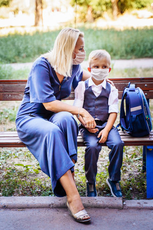 Mom and her son, a schoolboy, are sitting on a bench in the yard. They put on medical masks to protect themselvesの写真素材