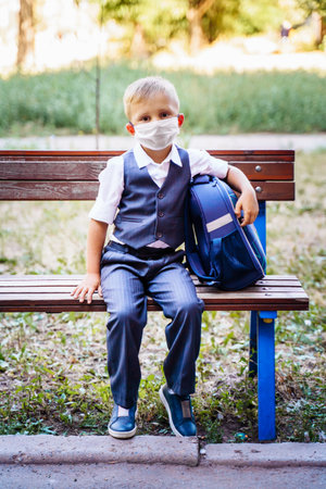 Young schoolboy in a protective medical mask sits on a bench in the park in front of the school. Dressed in formal uniform, holding backpack nearby. New reality during  pandemic.の写真素材