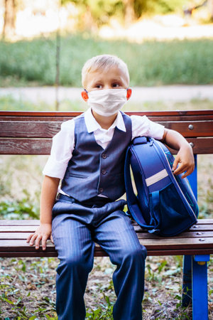 Young schoolboy in a protective medical mask sits on a bench in the park in front of the school. Dressed in formal uniform, holding backpack nearby. New reality during  pandemic.の写真素材