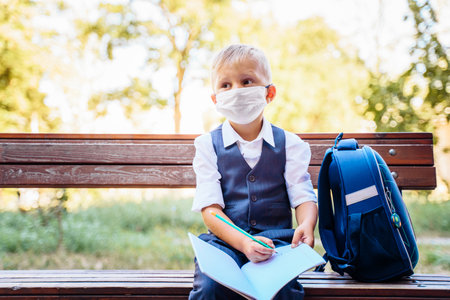 Elementary school student sits on a bench. Dressed in smart school uniform. Holding notebook on his knees and writing in it. New normal. Wears a medical protective maskの写真素材