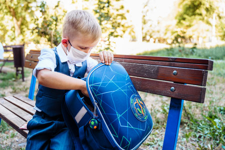 Young student of first grade stands in the schoolyard. Examines items from his backpack. Wearing protective medical mask due to new safety requirementsの写真素材