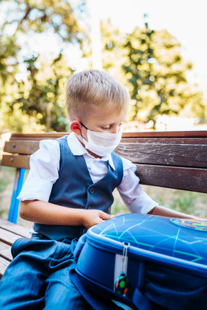 Young student of first grade stands in the schoolyard. Examines items from his backpack. Wearing protective medical mask due to new safety requirementsの写真素材
