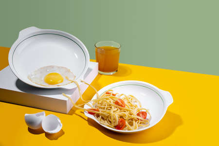 Concept pastafarianism. Monster noodles spagetti in a white plate attacking fried eggs for breakfast on bright yellow and a green background freshly squeezed orange juice and eggshellの写真素材