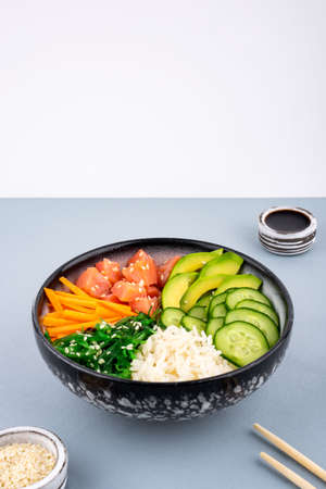 An overhead photo of poke bowl, traditional Hawaiian and Japanise raw fish salad, from seaweed, rice, salmon, cucumber, soy sause and avocado. with chopsticks and sesame seeds. Copy space, on a teal blue and white backgroundの写真素材
