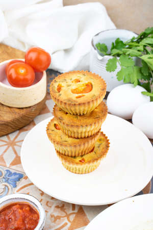 Small egg bite pastry muffins on a handmade white ceramic plate with a white linen napkin and egg, tomato, on a wooden board, and tile background. Finger food and mini food concept. Overhead. Top Viewの写真素材