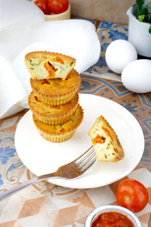 Small egg bite pastry muffins on a handmade white ceramic plate with a white linen napkin and egg, tomato, on a wooden board, and tile background. Finger food and mini food concept. Overhead. Top Viewの写真素材