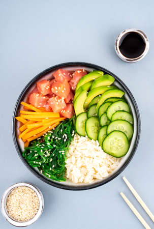 An overhead photo of poke bowl, traditional Hawaiian and Japanise raw fish salad, from seaweed, rice, salmon, cucumber, soy sause and avocado. with chopsticks and sesame seeds. Copy space, on a blue backgroundの写真素材