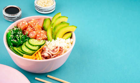 An overhead photo of poke bowl, traditional Hawaiian and Japanise raw fish salad, from seaweed, rice, salmon, cucumber, soy sause and avocado. with chopsticks and sesame seeds. Copy space, on a teal blue backgroundの写真素材