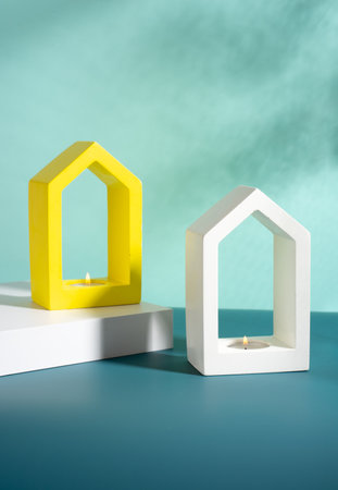 Creative composition. Still life. Shadows on the blue wall. Minimalistic house-shaped concrete candlestick white and yellow color with soy wax burning scented candleの写真素材