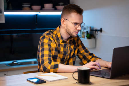 Portrait of male programmer in a yellow-black-gray shirt in a cage and glasses using Programs computer code in a Home. Works professionally on laptop. Concept programmer Day.の写真素材