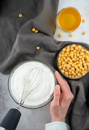 Chickpea aquafaba. Egg replacement. Vegan cooking concept. Men hands whippe whisk chickpeas liquid in glass bowl. chickpea in a ceramic handmade plate with linen napkin. On a gray background. Top viewの写真素材