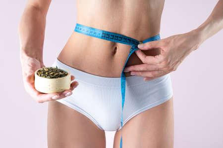Sports young woman tied a blue centimeter tape around her waist holding near the slender belly bowl of diet tea Genmaicha, fresh and dry leaves on pink background, closeup.Wellness and healthy conceptの写真素材