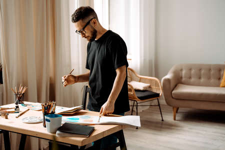 Portrait of concentrated male artist standing near a table drawing in a painting class in a studio,...