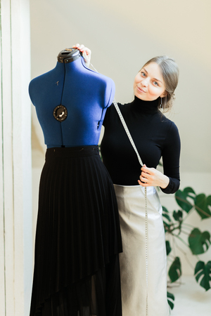 A young seamstress woman sets clothes on the mannequin. Portrait of seamstress and her hand close-up in the Studio. Focus on the mannequin with clothesの写真素材