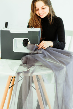 Young dressmaker woman sews clothes on sewing machine. Smiling seamstress and her hand close up in workshop. Focus on sewing machine and tissue.の写真素材