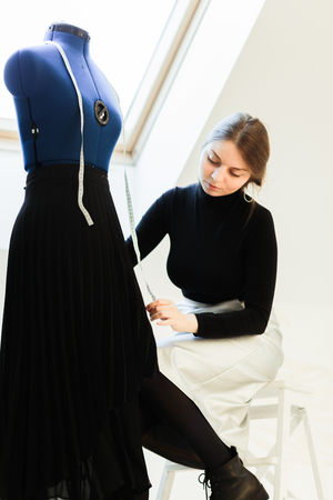 A young seamstress woman sets clothes on the mannequin. Portrait of seamstress and her hand close-up in the Studio. Focus on the mannequin with clothesの写真素材