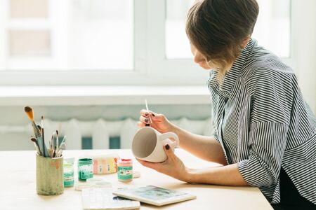 the pottery woman holds in hands a mug to paint the ornament, handmade ceramicの写真素材