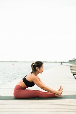 young asian girl doing yoga outdoors on the pier by the lake. she's wearing a black top and red...