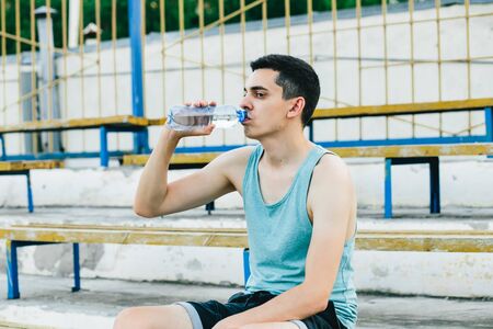 A young man sitting on a bench and tying his shoelaces in the fresh air in new York. He's wearing a t-shirt and gym shorts.の写真素材
