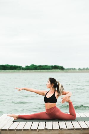 young asian girl doing yoga outdoors on the pier by the lake. she's wearing a black top and red...