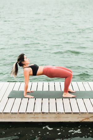Asian young girl doing yoga outdoors on the pier by the lake. she's wearing a black top and red...