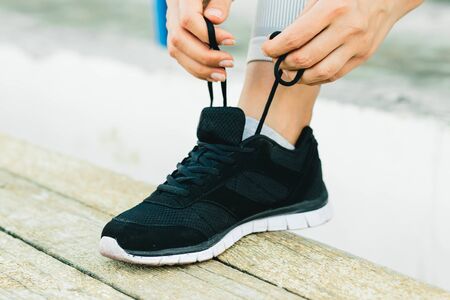 A young woman sitting on a bench and tying her shoelaces in the fresh air in new York. she's wearing...