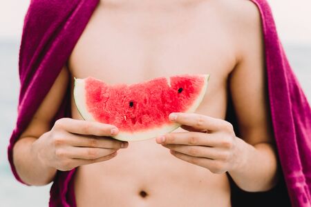 a young guy is standing near the sea and holding a slice of ripe watermelon