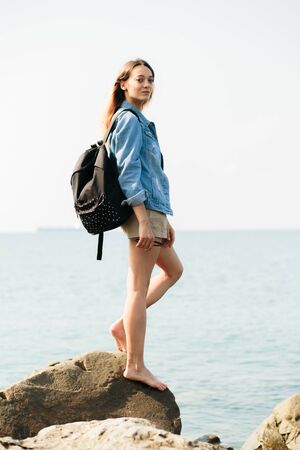 a young girl traveler with a backpack walks barefoot on large stones on the sea coastの写真素材