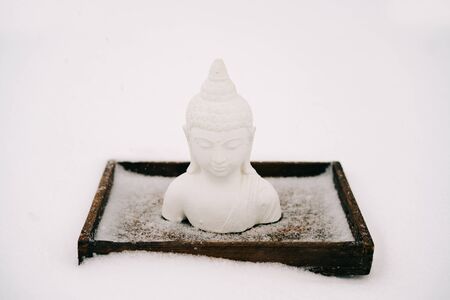 white Buddha figurine made of plaster on a wooden stand during a snowfallの写真素材