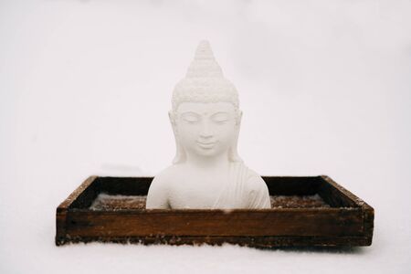 white Buddha figurine made of plaster on a wooden stand during a snowfallの写真素材