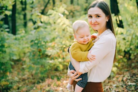 a young mother holds her little happy son in her arms among the autumn trees in the rays of the...