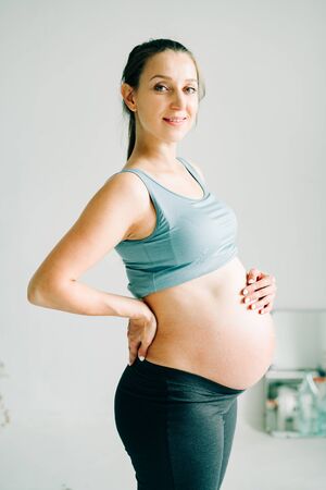 a young pregnant girl in sportswear is standing on a yoga Mat, smiling and holding her belly