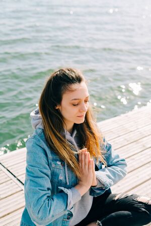 a young beautiful girl is sitting on a pier near the lake and doing meditation practiceの写真素材