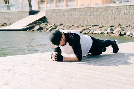 a young guy in sports black clothes on a pier near the lake doing sports exercises in the plank on a Sunny spring dayの写真素材