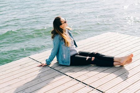 a young beautiful stylish girl is sitting on a pier near the lakeの写真素材