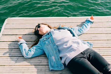 a young pretty girl in a denim jacket and glasses is lying on a pier near the lake